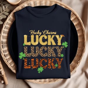 Peut inclure: T-shirt bleu marine avec l'inscription "Hucky Charms LUCKY" en paillettes dorées, motifs léopard et rayures tigrées. Des graphiques de trèfles verts sont également inclus. Le t-shirt est plié sur une assiette noire.