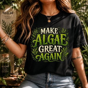 Peut inclure: Un t-shirt noir à manches courtes avec une impression graphique portant le texte "MAKE ALGAE GREAT AGAIN" en lettres stylisées vertes et crème, entouré d'illustrations d'algues vertes complexes et de volutes décoratives.