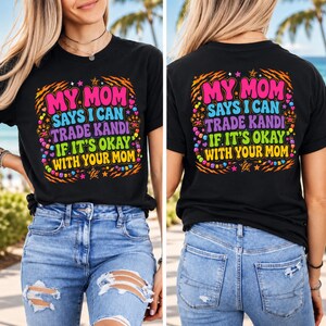 Peut inclure: Un t-shirt noir avec une impression graphique colorée portant le texte "MY MOM SAYS I CAN TRADE KANDI IF IT'S OKAY WITH YOUR MOM" entouré d'illustrations de perles, d'étoiles et de rayures de tigre en rose, bleu, vert et jaune.