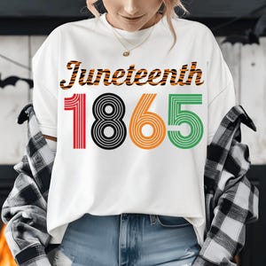 May include: White t-shirt featuring the word "Juneteenth" in orange and black tiger stripes. The number "1865" is printed below in red, black, orange, and green. The shirt is being worn.