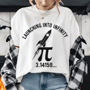 Peut inclure: T-shirt blanc avec l'inscription "LAUNCHING INTO INFINITY" en arc au-dessus d'un dessin de fusée noire. En dessous de la fusée, le symbole mathématique de pi, avec les chiffres 3.14159... imprimés en dessous.