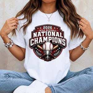 Op de afbeelding: Een wit T-shirt met korte mouwen en een grafisch ontwerp met de tekst 2026 NCAA BASEBALL NATIONAL CHAMPIONS. Het ontwerp bevat een honkbal, een trofee en twee gekruiste honkbalknuppels in het midden van een donkerrood en zwart schildembleem.