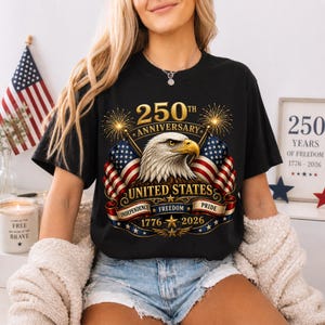 Peut inclure: T-shirt noir avec un motif patriotique célébrant le 250e anniversaire des États-Unis. Le graphisme comprend un aigle, des drapeaux américains et le texte "United States" et "Independence, Freedom, Pride 1776-2026".