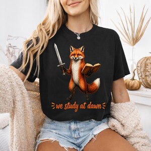 Peut inclure: T-shirt noir avec un dessin de renard tenant une épée et un livre. Le renard est orange et blanc. Le texte "we study at dawn" est imprimé en orange en dessous.
