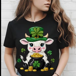 Peut inclure: T-shirt noir avec une vache de dessin animé portant un chapeau de leprechaun vert et tenant une marmite pleine de pièces d'or. La vache a un trèfle sur son chapeau. Des trèfles et des pièces d'or supplémentaires sont éparpillés.