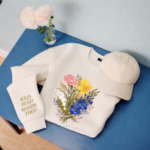 Peut inclure: Sweat-shirt crème avec un motif de bouquet floral composé de fleurs roses, jaunes et bleues. Une casquette de baseball crème assortie est posée à côté du sweat-shirt. La manche gauche affiche les noms "JULIA HUGO MANON THÉO" en or.