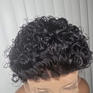 May include: A short, black wig featuring tight, wet-look curls and a sheer lace front base. The hair is styled in a cropped cut with defined ringlets that cover the top and sides, transitioning to a visible, fine mesh lace panel at the forehead.