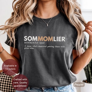 Puede incluir: Una camiseta gris oscuro con la palabra "SOMMOMLIER" en letras blancas y beige. La camiseta incluye la definición de la palabra y la frase "Una madre que domina el emparejamiento del caos con un gran vino."