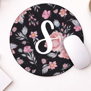Może przedstawiać: Okrągła czarna podkładka pod mysz z kwiatowym wzorem i białym znakiem "S". Kwiaty są różowe, a liście szare. Biała mysz komputerowa leży obok podkładki.