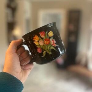 May include: Black speckled ceramic mug featuring a bouquet of tulips in shades of red, orange, and yellow. The mug has a black handle and a white speckled pattern. The floral design is painted on the front of the mug.