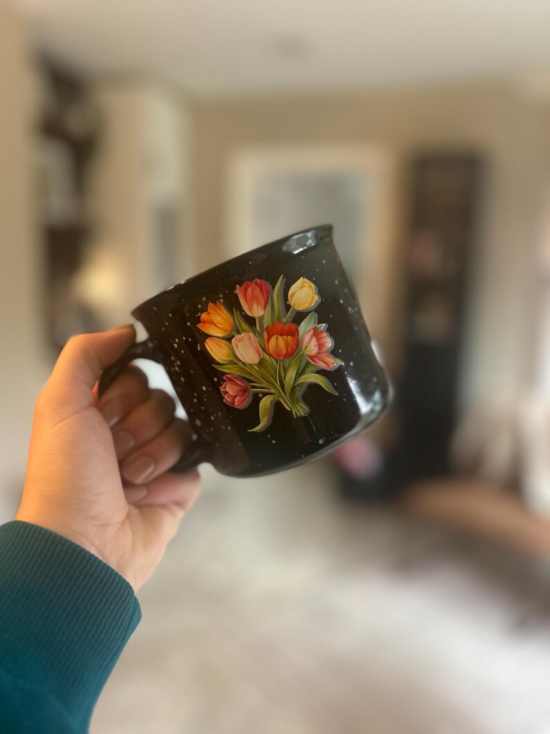 May include: Black speckled ceramic mug featuring a bouquet of tulips in shades of red, orange, and yellow. The mug has a black handle and a white speckled pattern. The floral design is painted on the front of the mug.