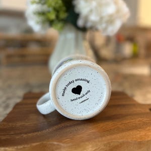 May include: White speckled ceramic mug with the words "make today amazing" and a black heart. The mug is on a wooden cutting board. The bottom of the mug also says "hand wash only".