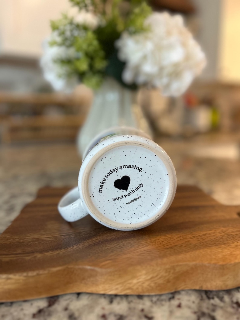 May include: White speckled ceramic mug with the words "make today amazing" and a black heart. The mug is on a wooden cutting board. The bottom of the mug also says "hand wash only".