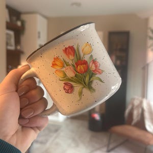 May include: White ceramic mug with a speckled finish and a black rim. The mug features a bouquet of tulips in shades of red, orange, and yellow. The mug has a curved handle.