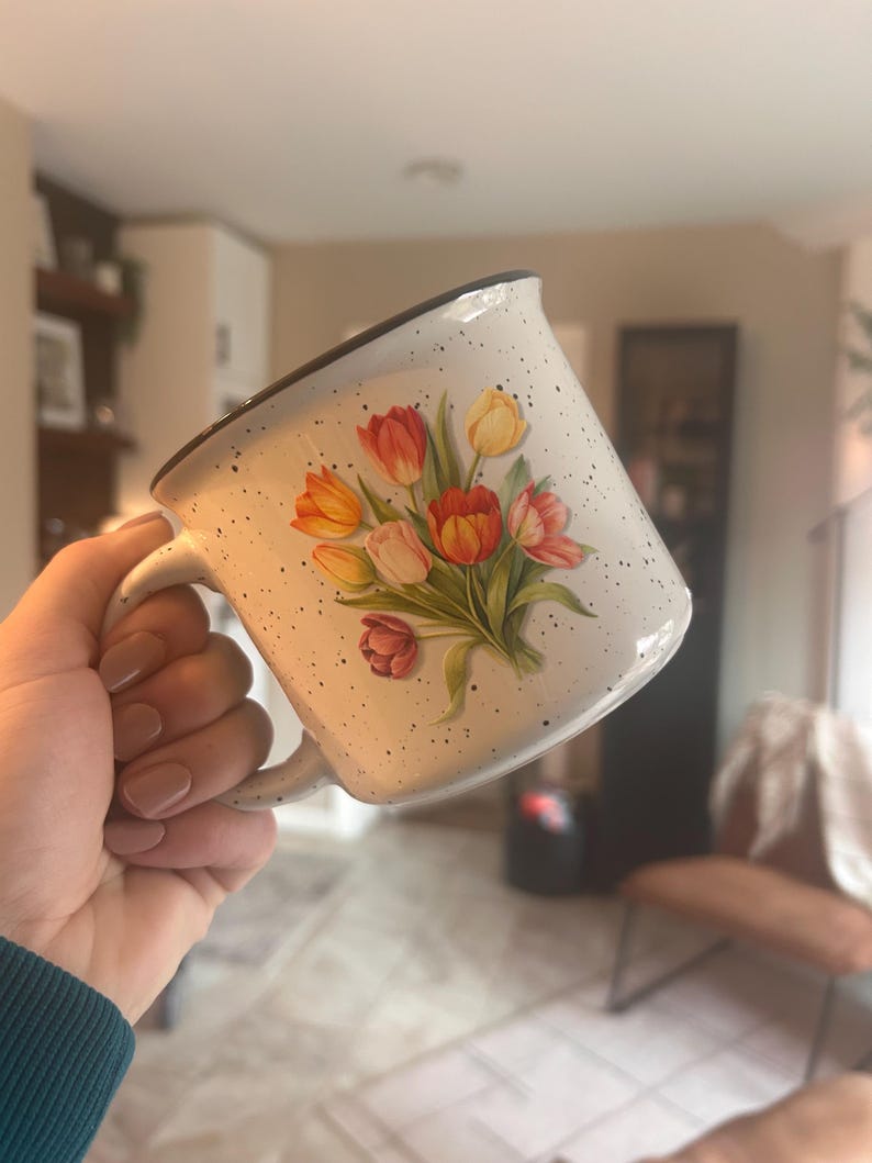 May include: White ceramic mug with a speckled finish and a black rim. The mug features a bouquet of tulips in shades of red, orange, and yellow. The mug has a curved handle.