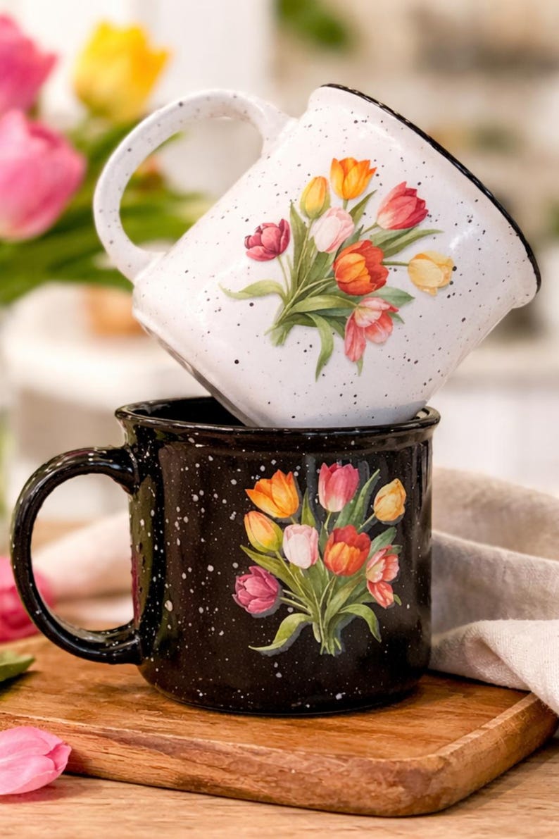May include: Two ceramic mugs, one white and one black, stacked on a wooden surface. Both mugs feature a floral design of colorful tulips. The white mug has a speckled pattern, and the black mug has a speckled pattern as well.