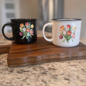 May include: Two ceramic mugs with tulip designs. One mug is black with white speckles, the other is white with speckles. Both mugs have a black rim and a floral bouquet of red, orange, and yellow tulips. They sit on a wooden cutting board.