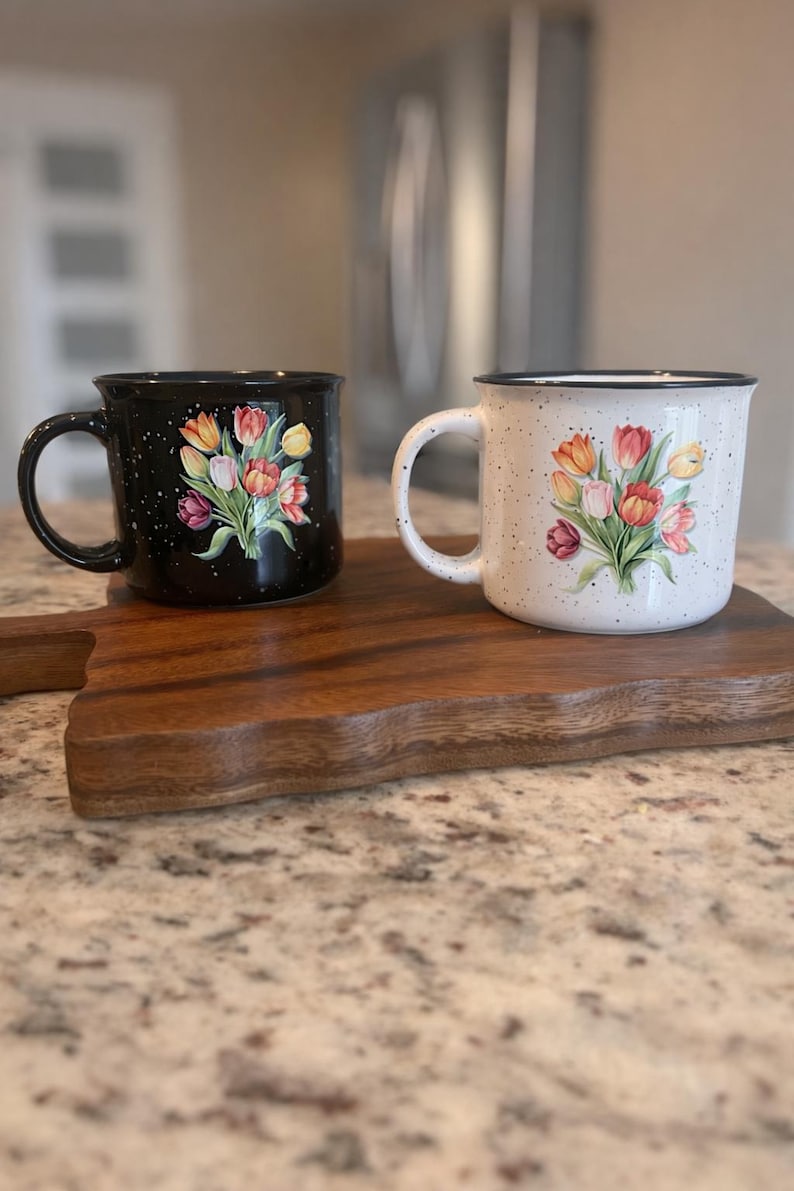 May include: Two ceramic mugs with tulip designs. One mug is black with white speckles, the other is white with speckles. Both mugs have a black rim and a floral bouquet of red, orange, and yellow tulips. They sit on a wooden cutting board.