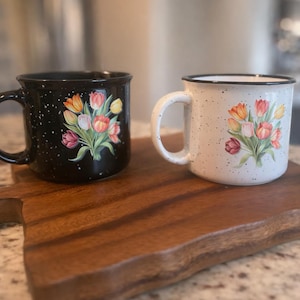 May include: Two ceramic mugs with floral designs. One mug is black with white speckles, the other is white with black speckles. Both mugs feature a bouquet of tulips in shades of orange, pink, and yellow. The mugs are on a wooden cutting board.