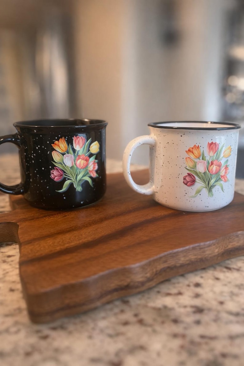 May include: Two ceramic mugs with floral designs. One mug is black with white speckles, the other is white with black speckles. Both mugs feature a bouquet of tulips in shades of orange, pink, and yellow. The mugs are on a wooden cutting board.
