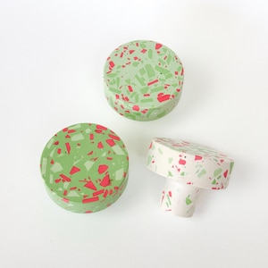 May include: Three bottle stoppers with a terrazzo design. The stoppers are round and cylindrical, with a light green base and red and green flecks. One stopper is upright, and two are lying flat.