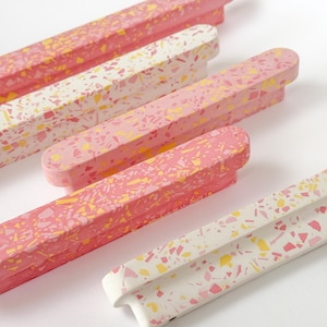 Op de afbeelding: Een verzameling rechthoekige terrazzo-stijl meubelgrepen in roze, wit en geel. De grepen hebben een afgeronde bovenkant en een kleine, gebogen handgreep. Het terrazzo-patroon bestaat uit kleine, onregelmatige vormen.