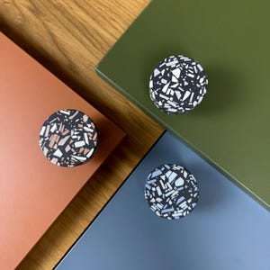 Puede incluir: Tres pomos redondos para armarios con diseño terrazo. Cada pomo presenta una base negra con un mosaico de virutas blancas, grises y de colores. Los pomos están colocados sobre superficies de color naranja, verde y azul.