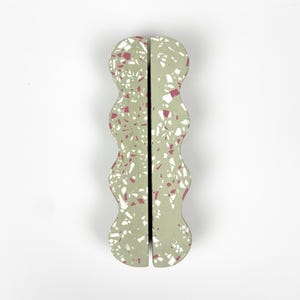 May include: Two-piece, wavy-edged decorative object with a terrazzo design. The object is light green with white and maroon flecks. The pieces are separated by a thin black line, and the background is white.