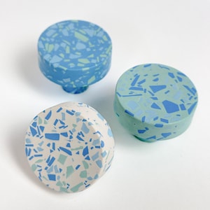 May include: Three decorative cabinet knobs with terrazzo-style designs. The knobs are in shades of blue, green, and white. The knobs are round with a cylindrical base. The terrazzo pattern features irregular shapes in varying shades.