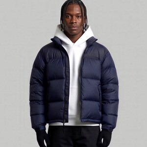 Puede incluir: Una chaqueta acolchada de color azul marino con canales acolchados horizontales y una cremallera frontal completa, usada sobre una sudadera con capucha blanca lisa. La chaqueta presenta un cuello alto y mangas largas con puños ajustables.