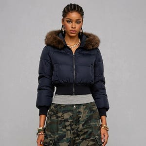 May include: A navy blue cropped puffer jacket featuring a brown faux fur-lined hood, a front zipper closure, and black ribbed cuffs and hem. The jacket is worn over a grey shirt and camouflage print cargo pants with visible pockets and belt loops.