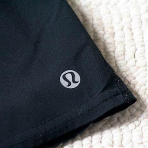 May include: Close-up of a black athletic short with a silver logo on the corner. The shorts are made of a smooth, dark fabric and have a stitched hem. The logo is a stylized 'L' shape.