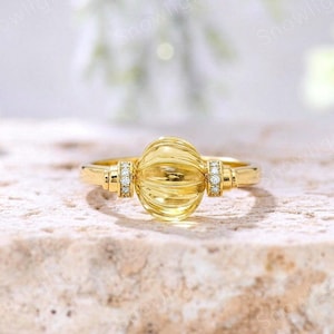Può includere: Un anello in oro con una pietra preziosa gialla centrale, sferica e scanalata. La fascia è decorata con piccole pietre trasparenti a taglio rotondo su entrambi i lati dell'elemento centrale. Il metallo ha una finitura lucida.