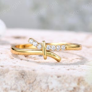 May include: A gold-colored ring with a unique design. The ring features a band with a curved accent and a vertical bar, adorned with small, clear gemstones. The ring is set against a neutral background, highlighting its elegant design.