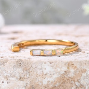 May include: A gold ring featuring three rectangular opal stones. The ring has an open design, with the ends of the band not connected. The ring is displayed on a textured, light-coloured surface. The ring's design is modern.