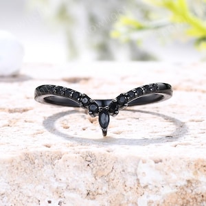 May include: A black V-shaped ring featuring a series of small, round black stones set along the band, with a single marquise-cut black stone positioned at the center point of the chevron design, creating a symmetrical and pointed aesthetic.