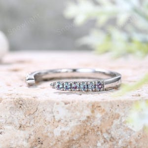 May include: A silver ring with a row of small, colorful gemstones. The ring is set on a textured, light-colored surface. The gemstones are a mix of blue and purple hues. The ring has a modern, open design.