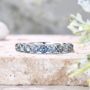 Op de afbeelding: Een zilveren ring met een rij ronde, lichtblauwe edelstenen. De edelstenen variëren in tint, waardoor een gradiënteffect ontstaat van licht naar donkerder blauw en groen. De ring is geplaatst tegen een neutrale, getextureerde achtergrond.