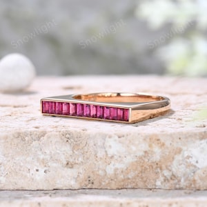 May include: A rose gold ring with a rectangular top set with a row of rectangular, vibrant pink gemstones. The ring is displayed on a textured, light-colored surface.