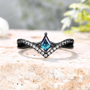 May include: A black metal ring featuring a kite-shaped, color-shifting blue and purple center stone. The band is designed in a chevron shape and is accented with a row of small, clear round stones along the top edge of the V-shaped setting.
