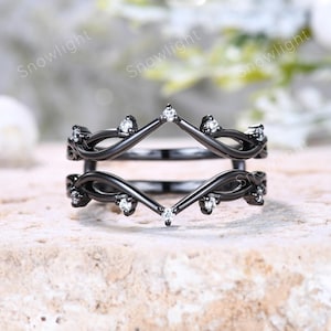 May include: A black metal ring guard featuring a double band design with a pointed chevron shape. The ring is accented with small, clear, round-cut stones set along the top and bottom edges of the frame, creating a symmetrical, open-work look.