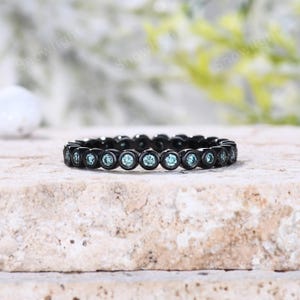 May include: A black metal eternity band featuring a continuous row of round, light blue stones set in individual bezels. The ring displays a uniform design with each stone securely encased in a dark, matte-finished metal frame around the band.