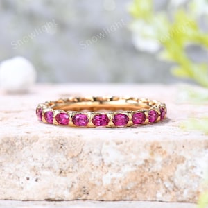 May include: A gold ring with a band of oval-cut, vibrant pink gemstones. The ring is set on a light-colored, textured surface, with soft, blurred greenery in the background. The word "Snowlight" is visible in the image.