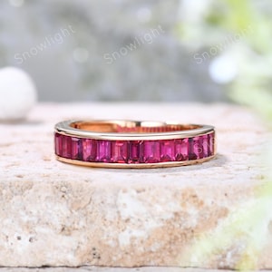May include: A rose gold ring featuring a band of rectangular, vibrant pink gemstones. The ring is set on a neutral-toned surface, with a soft, blurred background. The gemstones are closely set, creating a continuous line of color.