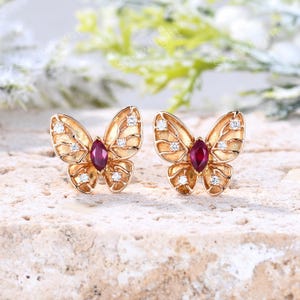 May include: A pair of gold butterfly-shaped stud earrings featuring a central marquise-cut deep red gemstone and four small round clear accent stones on each wing. The textured gold metal wings have a detailed vein-like pattern throughout.