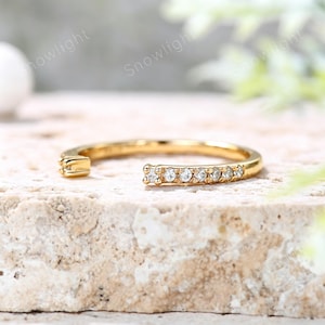 Könnte beinhalten: Ein goldfarbener Ring mit einer Reihe kleiner, klarer Edelsteine. Der Ring hat ein offenes Design mit einem kleinen, dekorativen Detail an einem Ende. Der Ring wird auf einer hellen, strukturierten Oberfläche gezeigt.