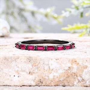 May include: A thin black metal band ring featuring a continuous row of vibrant red baguette-cut gemstones set horizontally. The stones are arranged in a channel setting, creating a sleek and uniform look across the entire circumference of the ring.
