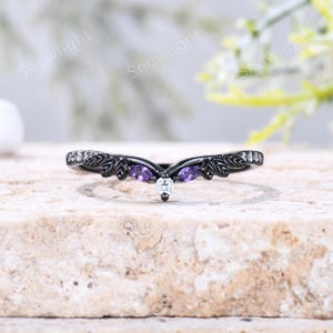 May include: A black metal V-shaped ring featuring a central round white stone, two marquise-cut purple stones, and small clear stones along the band. The band is detailed with a leaf-like texture on both sides of the center stones.