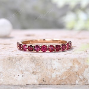 May include: A rose gold ring featuring a band of vibrant red gemstones. The gemstones are evenly spaced around the ring, creating a continuous line of color. The ring is set against a neutral background, highlighting its design.