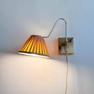 Könnte beinhalten: Eine Wandleuchte mit einem plissierten, konisch geformten Schirm in Gelb- und Brauntönen. Die Lampe hat einen Metallarm und eine quadratische Basis. Das Kabel ist sichtbar und hängt herunter. Die Lampe ist eingeschaltet und strahlt ein warmes Licht aus.